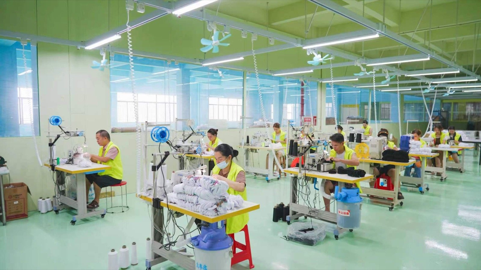 Workers operate sewing equipment beside stacks of socks in a production room.